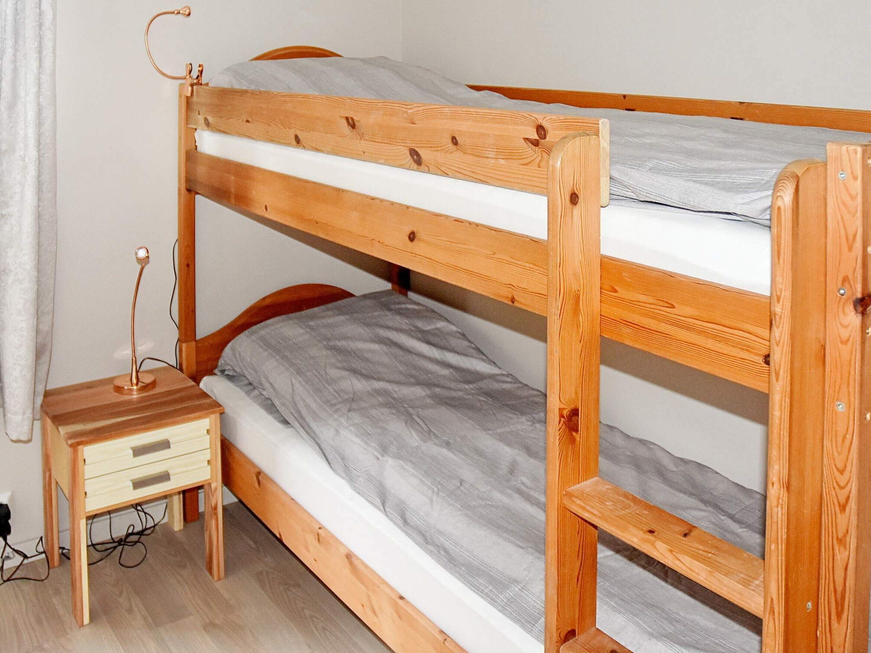 A bunk bed with a wooden frame, a bedside table with drawers, and a lamp.