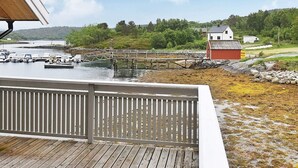 A wooden deck with a railing overlooking a dock area with boats, a small red building, and a forested hill in the background.