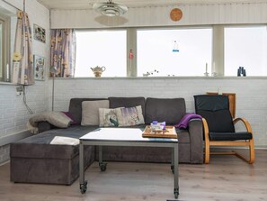 A living room with a sectional sofa, a small table with a tray, a black chair, and a window with a view of the sky. - 3 Person Holiday Home in Grasten (Grasten)