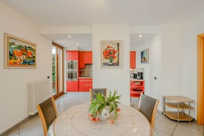 A dining area with a round table, chairs, a vase with flowers, and a kitchen in the background.