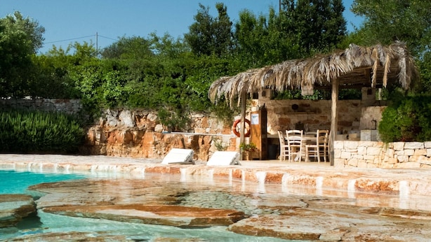 Ein klares Schwimmbecken mit natürlichen Felsen, einer Holzterrasse und einem Gebäude mit Strohdach.
