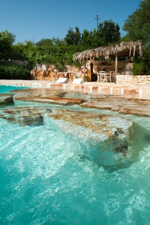 Ein klares Schwimmbecken mit natürlichen Felsen, einer Holzterrasse und einem Gebäude mit Strohdach.
