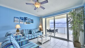 A room with a blue sofa set, a white coffee table, and a ceiling fan. There are large windows and a view of the sky. - 14th Floor 3BR 2BA at the Boardwalk!! (Panama City)