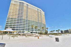 A multi-story hotel with balconies, a beach in the foreground, and palm trees.
