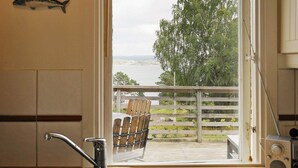 Eine Küche mit Spüle, ein Holzterrasse mit Stühlen und Blick auf ein Gewässer und Bäume.