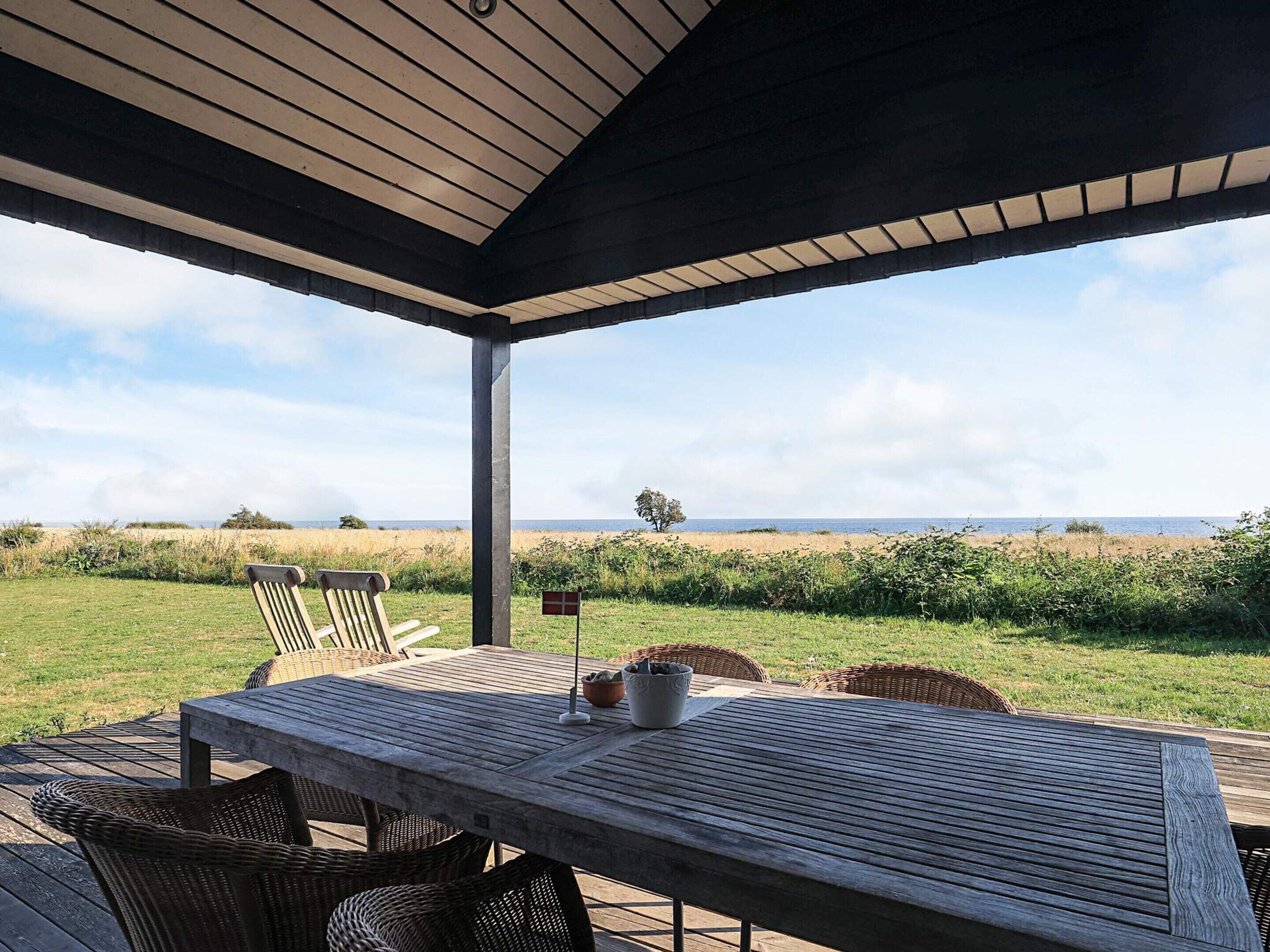 A covered outdoor seating area with a table and chairs, overlooking a grassy field and the sea.