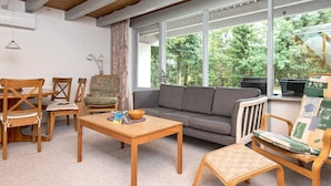 A cozy living room with a wooden coffee table, a grey sofa, and wooden chairs. Large windows offer a view of greenery outside. - 5 Person Holiday Home in Ebeltoft (Ebeltoft)