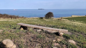 A wooden bench in a grassy area with a view of the sea and a distant island. - 5 Person Holiday Home in Ebeltoft (Ebeltoft)
