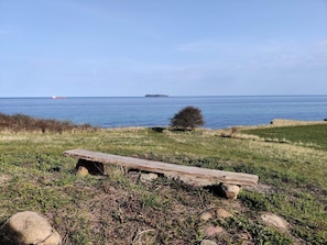 A wooden bench in a grassy area with a view of the sea and a distant island. - 5 Person Holiday Home in Ebeltoft (Ebeltoft)