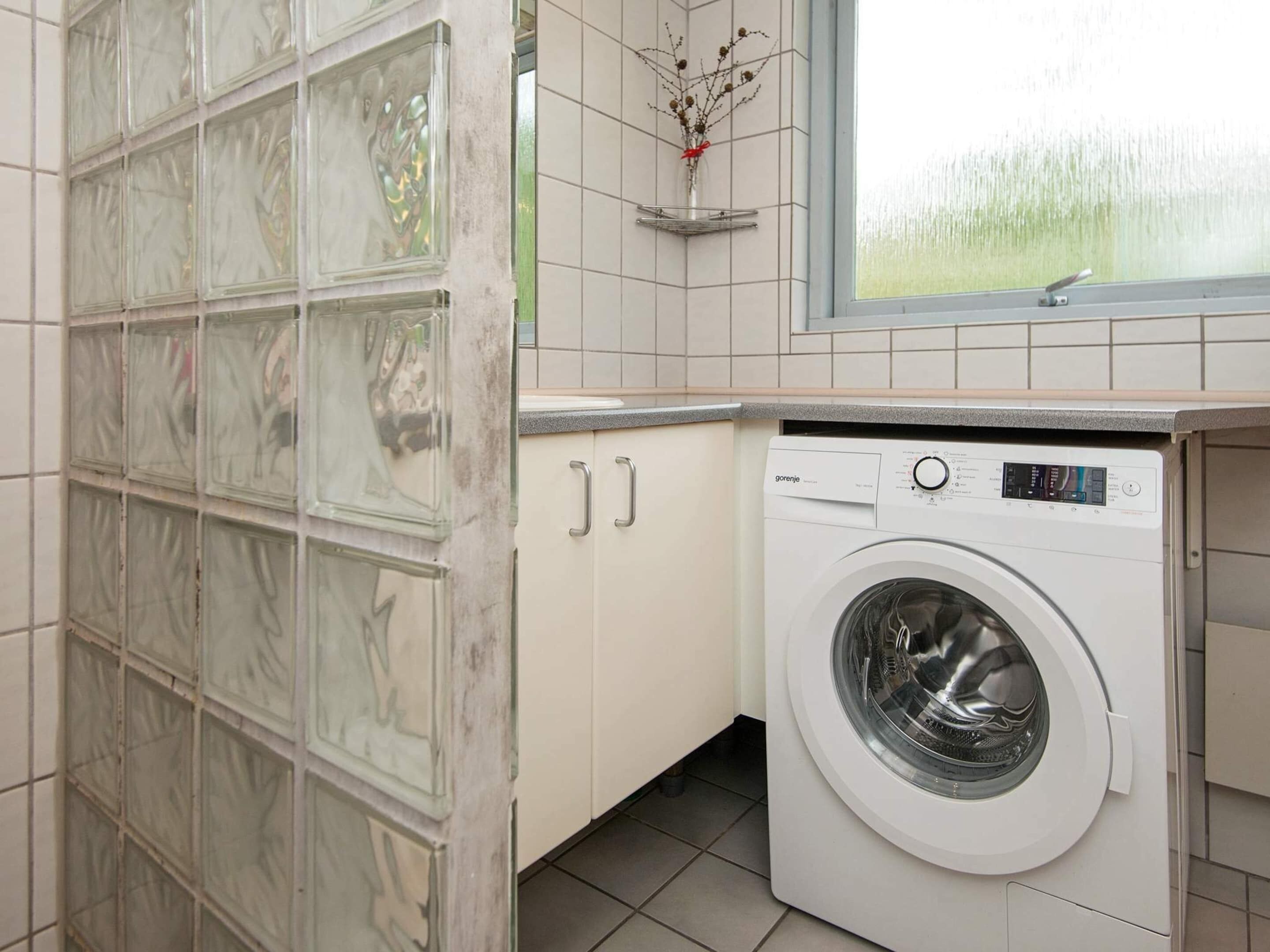A compact laundry room with a front-loading washing machine, a tiled backsplash, and a window with a view of greenery.