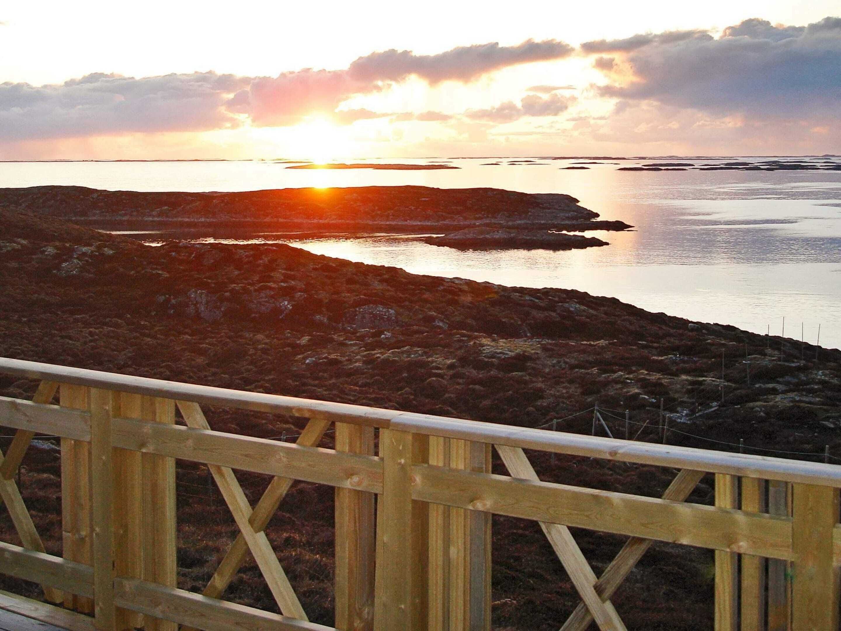 Una terraza de madera con vista a una costa rocosa, mientras el sol se pone sobre el agua.