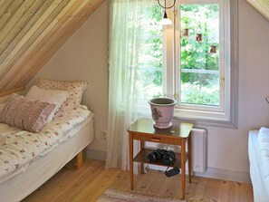A cozy bedroom with a sloped ceiling, a bed with floral bedding, a window with a view of greenery, a small table with a vase and a camera, and a radiator.