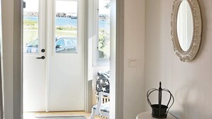 A room with a white door leading to a view of a car and a body of water. There is a white wall with a decorative mirror and a small black object on a white surface. - Holiday Home in Sölvesborg (Sölvesborg)