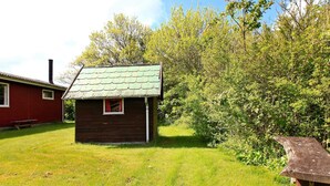 A small wooden cabin with a green roof, surrounded by lush greenery and a red building in the background.