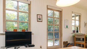 A room with a wooden table, a television on a white cabinet, and large windows with a view of greenery outside. - Stylish Holiday Home in Hals near Sea (Hals)
