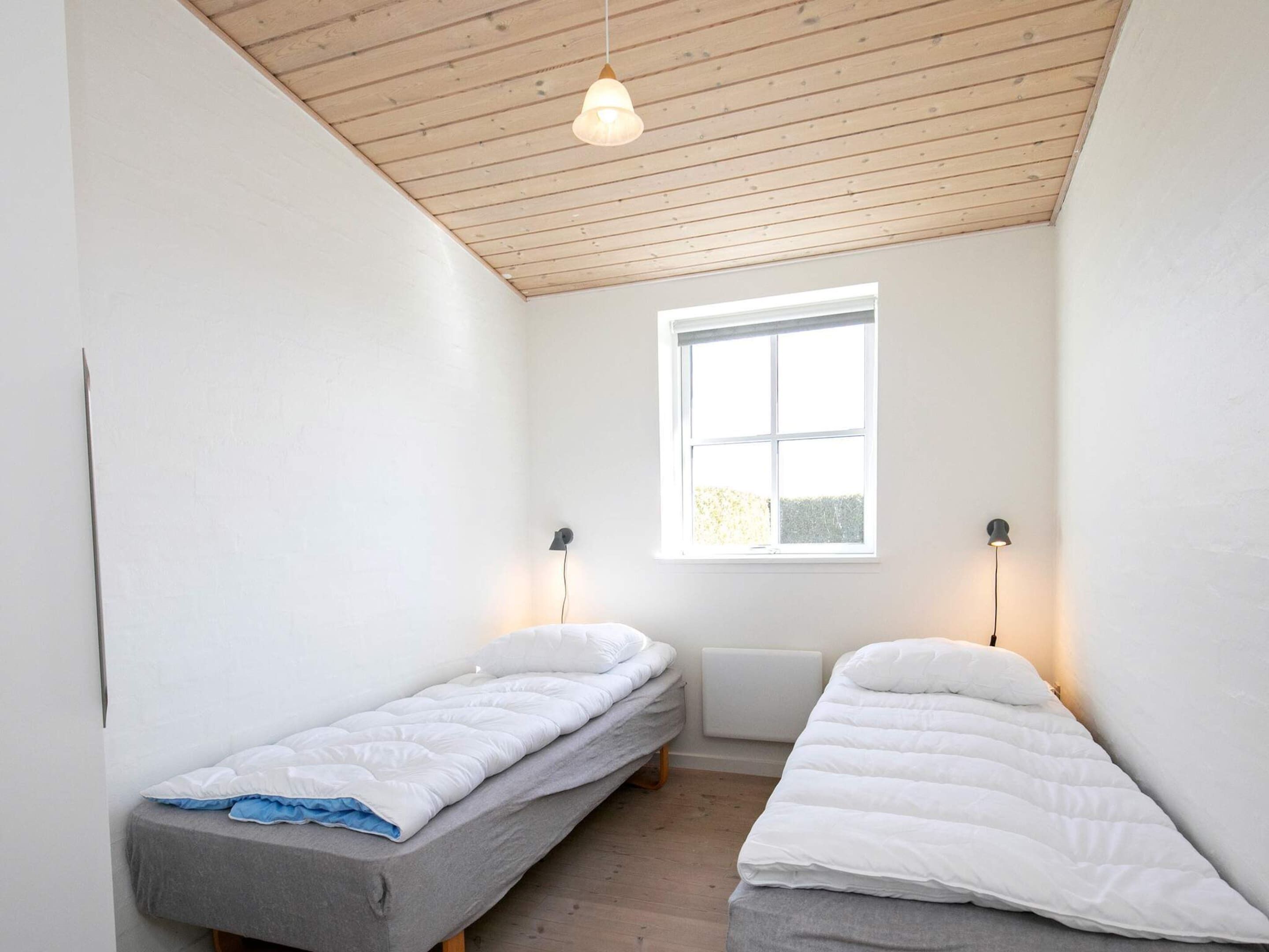 A small, bright room with two single beds, a window, and a wooden ceiling.