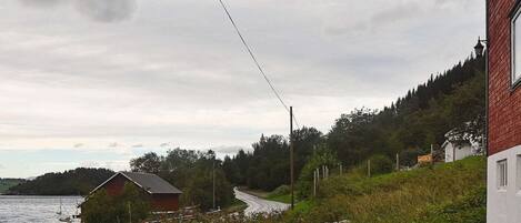 A coastal road with a house on the right, a boat near the water, and a forested hill in the background.
