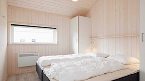 A bedroom with wooden panel walls, a bed with white bedding, a window, and a radiator. - Holiday Home in Großenbrode (Grossenbrode)