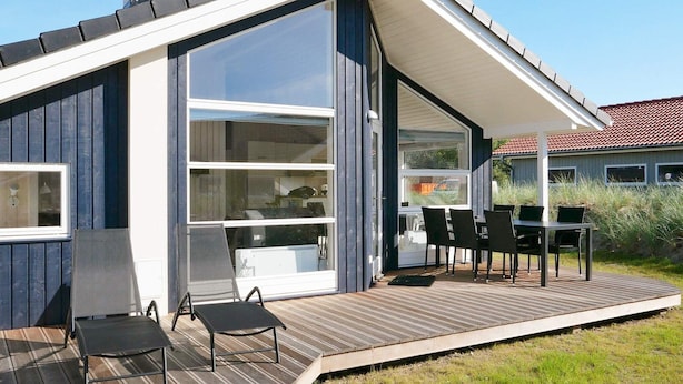 A modern house with a large glass door, a deck with outdoor furniture, and a clear view of the surrounding area. - Holiday Home in Großenbrode (Grossenbrode)