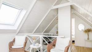 Acogedor dormitorio en el ático con paredes de paneles blancos, techo inclinado y tragaluz. Hay dos sillas de mimbre con cojines blancos y una pequeña mesa blanca con un jarrón y una vela.