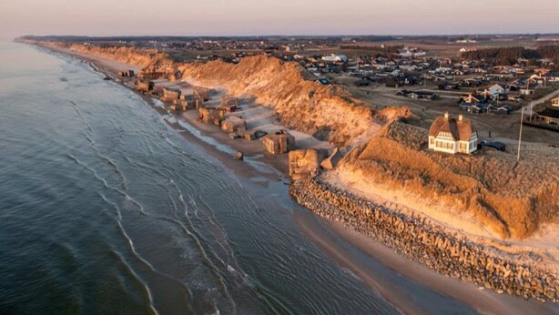 Un pueblo costero con un edificio destacado en una zona elevada, una playa con rocas y una vista distante de casas y campos.