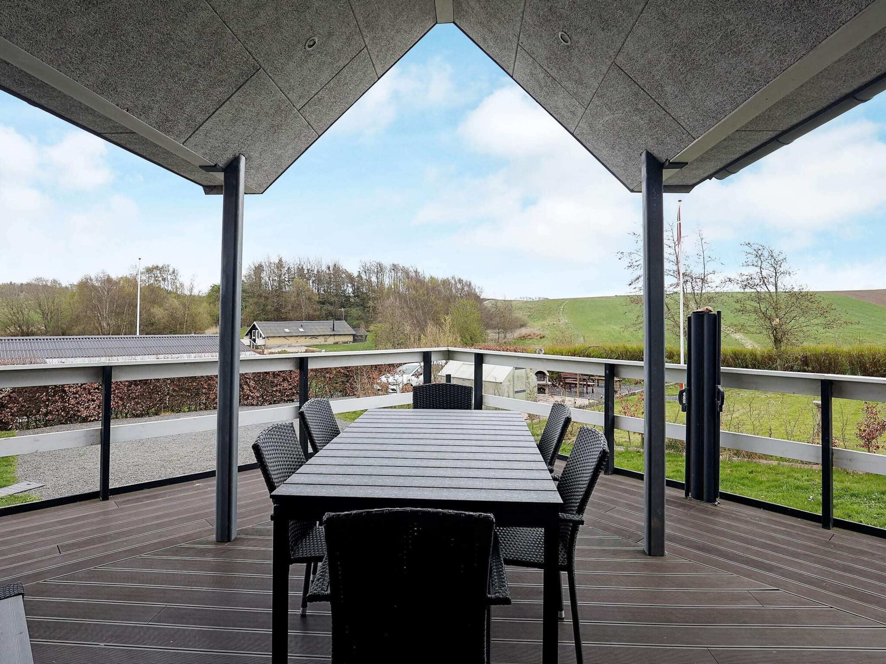 Een overdekt terras met een tafel en stoelen, uitzicht op het landschap, en een gebouw op de achtergrond.