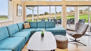 Un salon moderne avec un canapé d’angle, une table basse et un fauteuil gris. De grandes fenêtres offrent une vue sur l’extérieur.