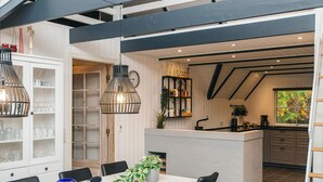 A modern kitchen with a central island, black chairs, a white dining table, and a hanging light fixture. - 10 Person Holiday Home in Blavand (Blavand)