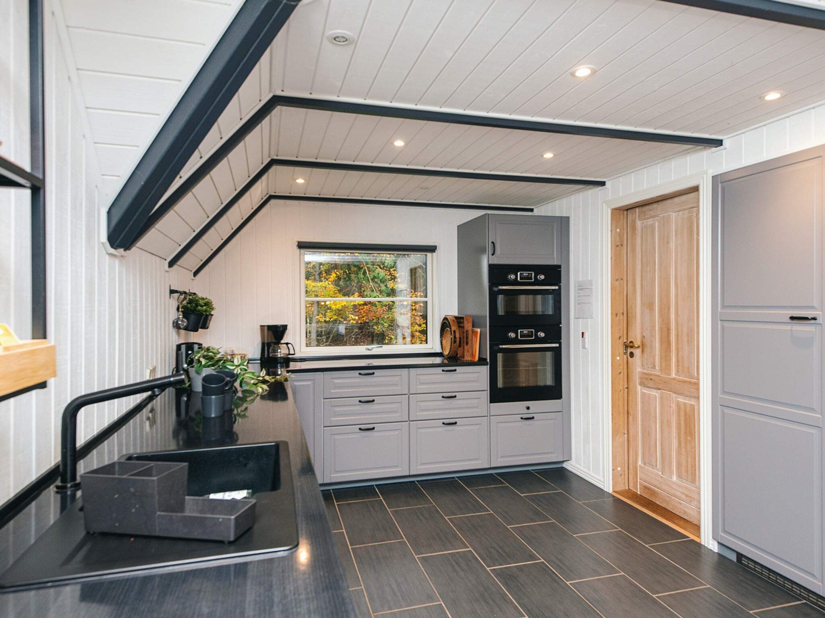 A modern kitchen with a built-in oven, a window with a view of trees, and a wooden door.
