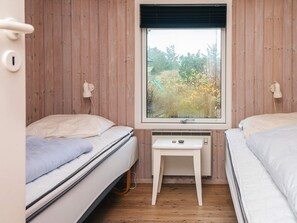 A bedroom with two beds, a small table, a window with a view of greenery, and a wall-mounted heater.