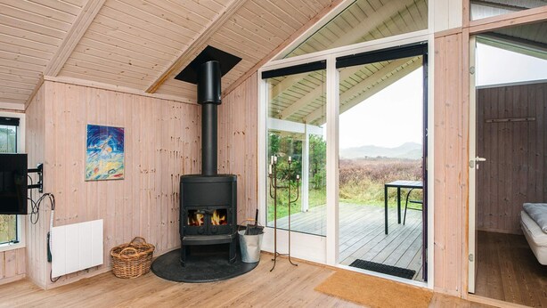 A cozy room with a wood-burning stove, wooden walls, a large sliding glass door leading to an outdoor deck, and a painting on the wall. - Serene Holiday Home in Oksbøl near Beach (Oksbol)