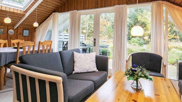 A cozy room with a wooden ceiling, a grey sofa, a wooden table, and a view of the outdoors through large windows.