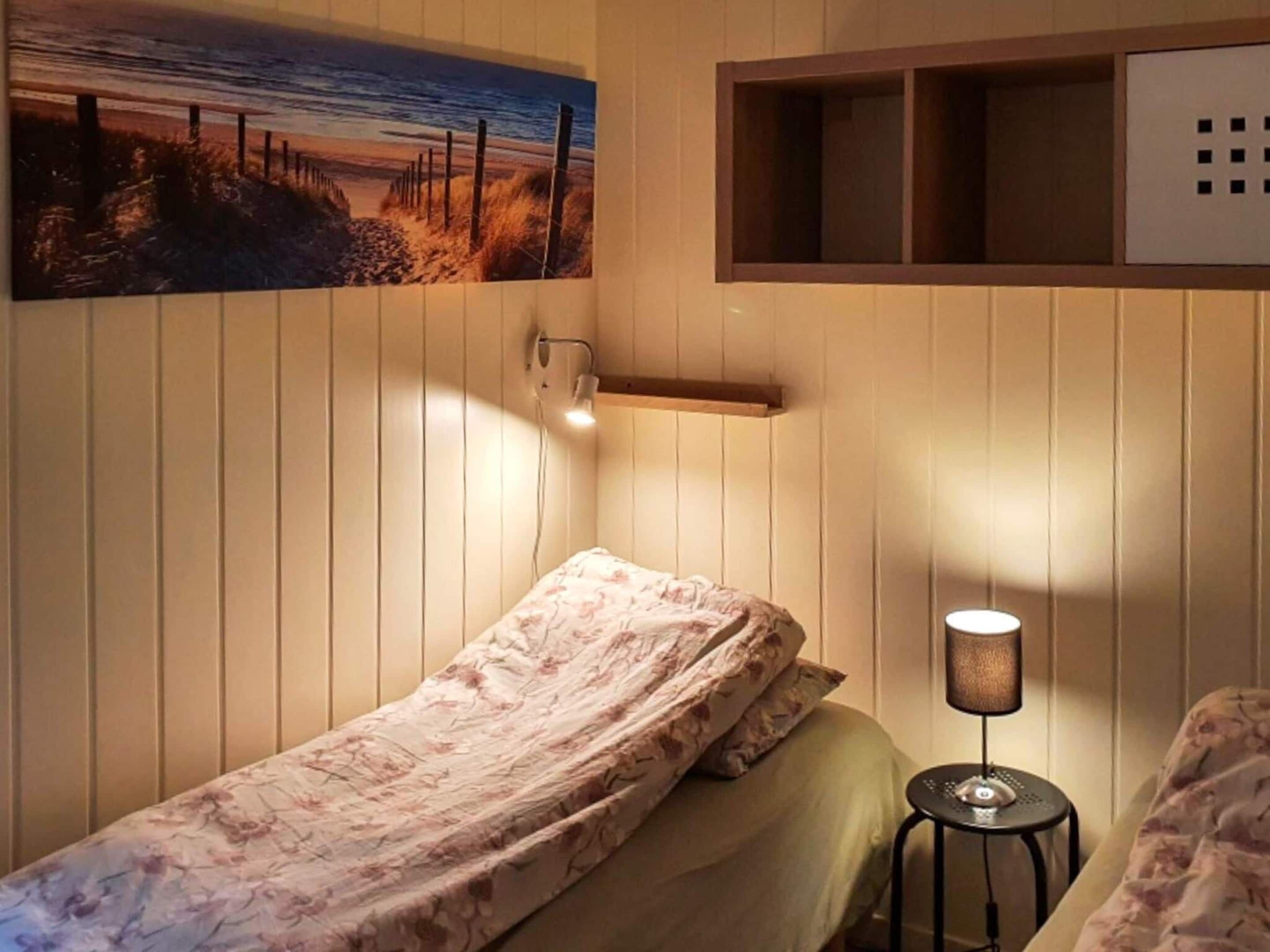 Een enkel bed met een bloemenbedekker, een nachtkastje met een lamp, een wandplank en een schilderij van een strandgezicht.