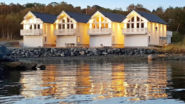 A waterfront property with multiple connected buildings, a stone retaining wall, and a calm body of water reflecting the structures. - Holiday Home in BrattvÃ¥g (Ålesund)