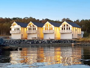 A waterfront property with multiple connected buildings, a stone retaining wall, and a calm body of water reflecting the structures. - Holiday Home in BrattvÃ¥g (Ålesund)