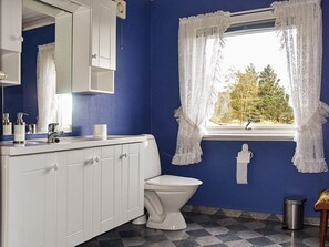 Ein Badezimmer mit blauer Wand, weißen Schränken, einer weißen Toilette, einem Fenster mit weißen Vorhängen und einem Metall-Mülleimer.