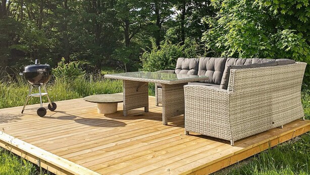 Eine Holzterrasse mit Korbmöbeln, darunter eine Sitzecke und ein Tisch, sowie ein Grill.