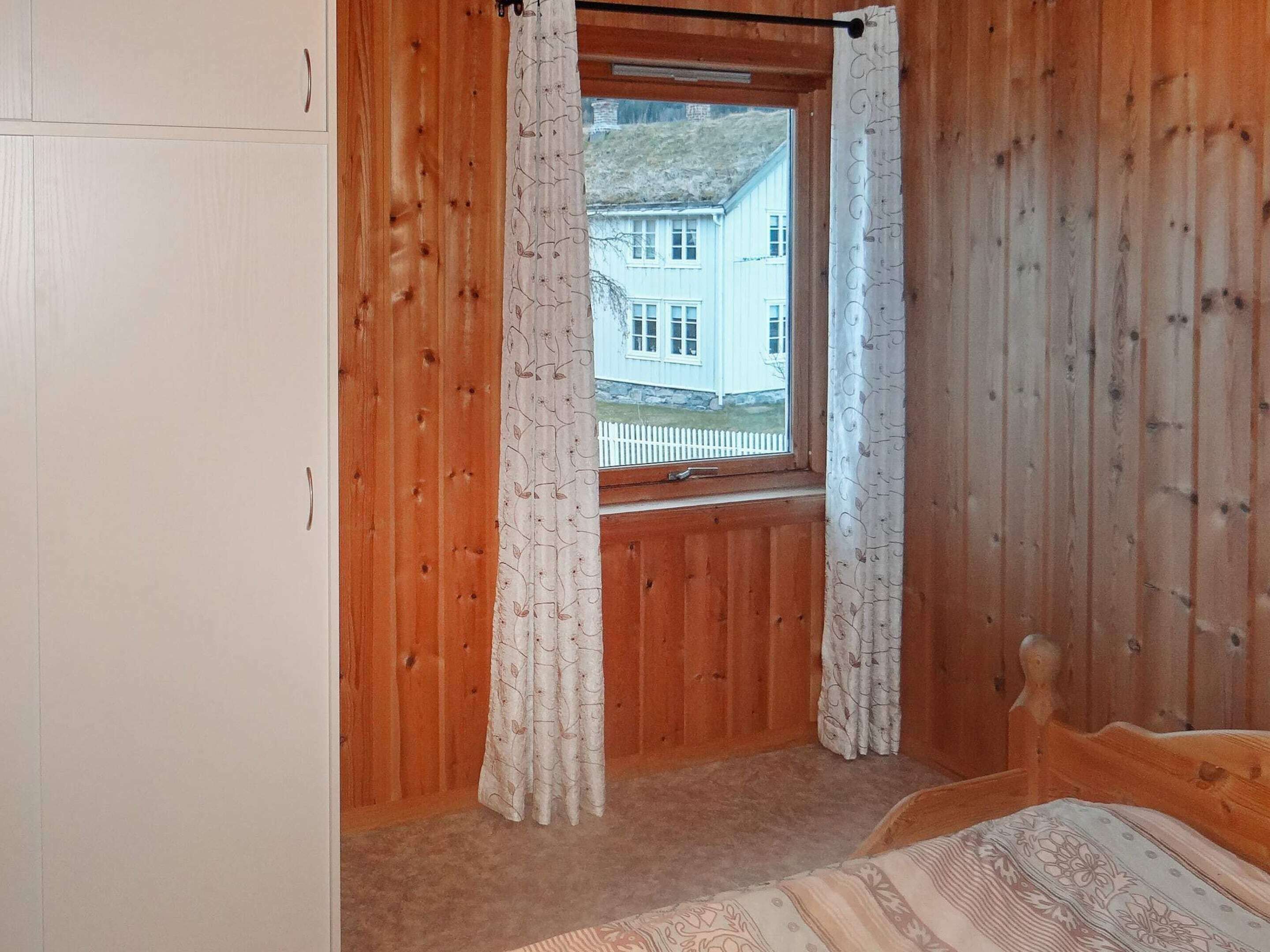 Makuuhuone, jonka seinät ovat puuta, ikkunassa on verhot, sängyssä on kuviollinen päiväpeite, ja ikkunasta näkyy valkoinen rakennus.