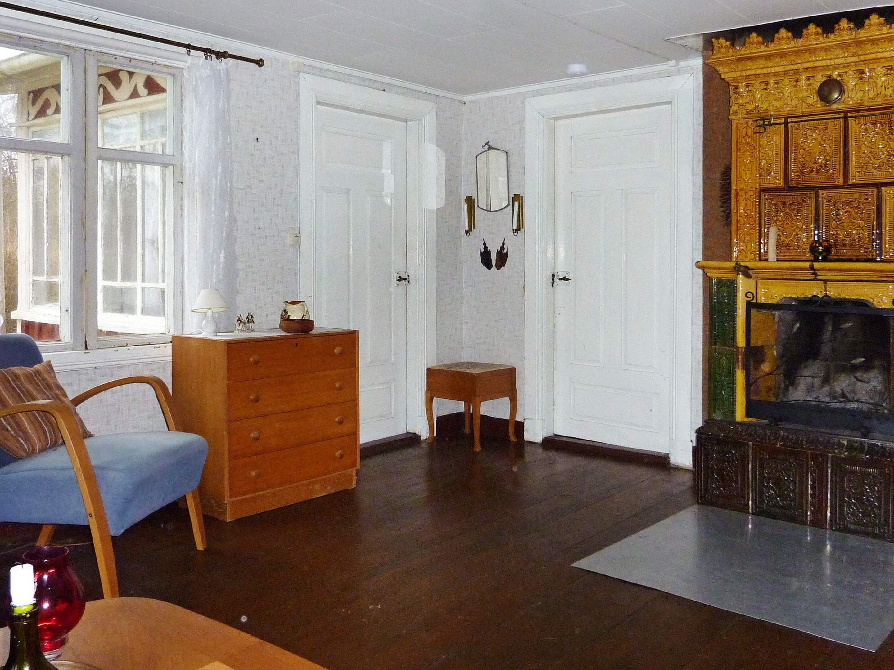 A room with a wooden dresser, a blue chair, a small table, and a fireplace with intricate carvings.