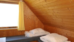 A small, cozy room with wooden walls, a sloped ceiling, a window with yellow curtains, and a bed with white bedding.
