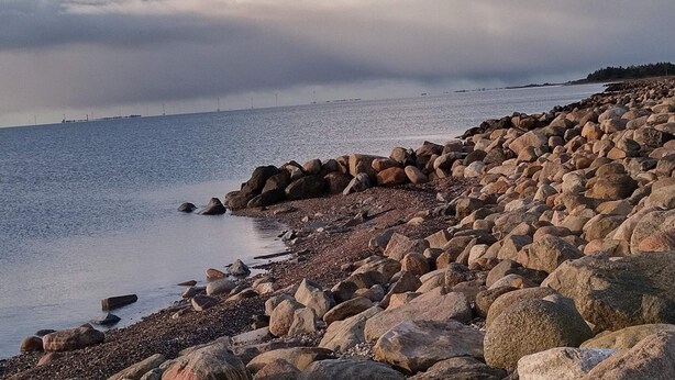 A rocky shoreline with large boulders and pebbles, leading to calm water.