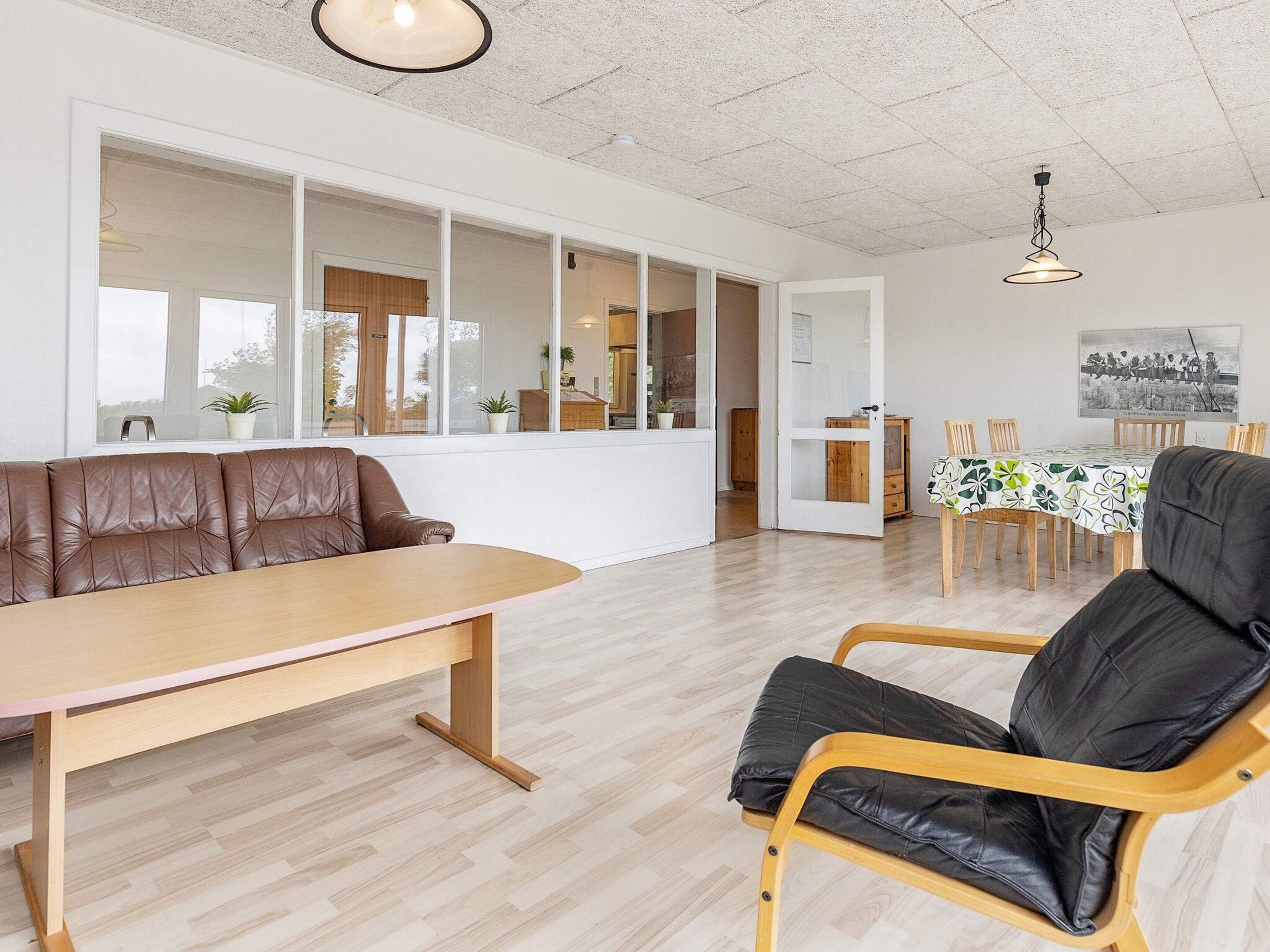 A modern living room with a sofa, a dining table, and a chair. There is a kitchen area visible through a glass door.