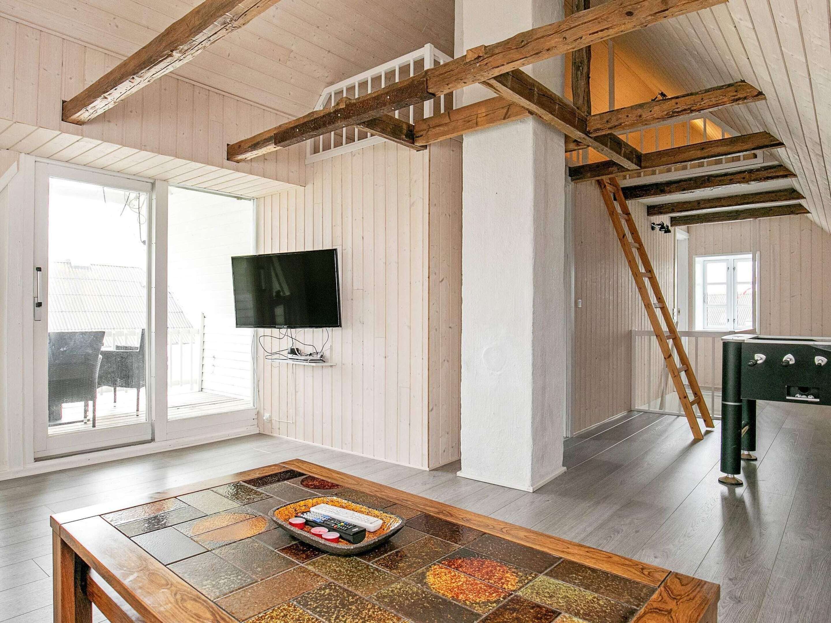Um quarto amplo com teto de madeira, uma televisão na parede, uma mesa de jantar com um tabuleiro de condimentos e uma escada.