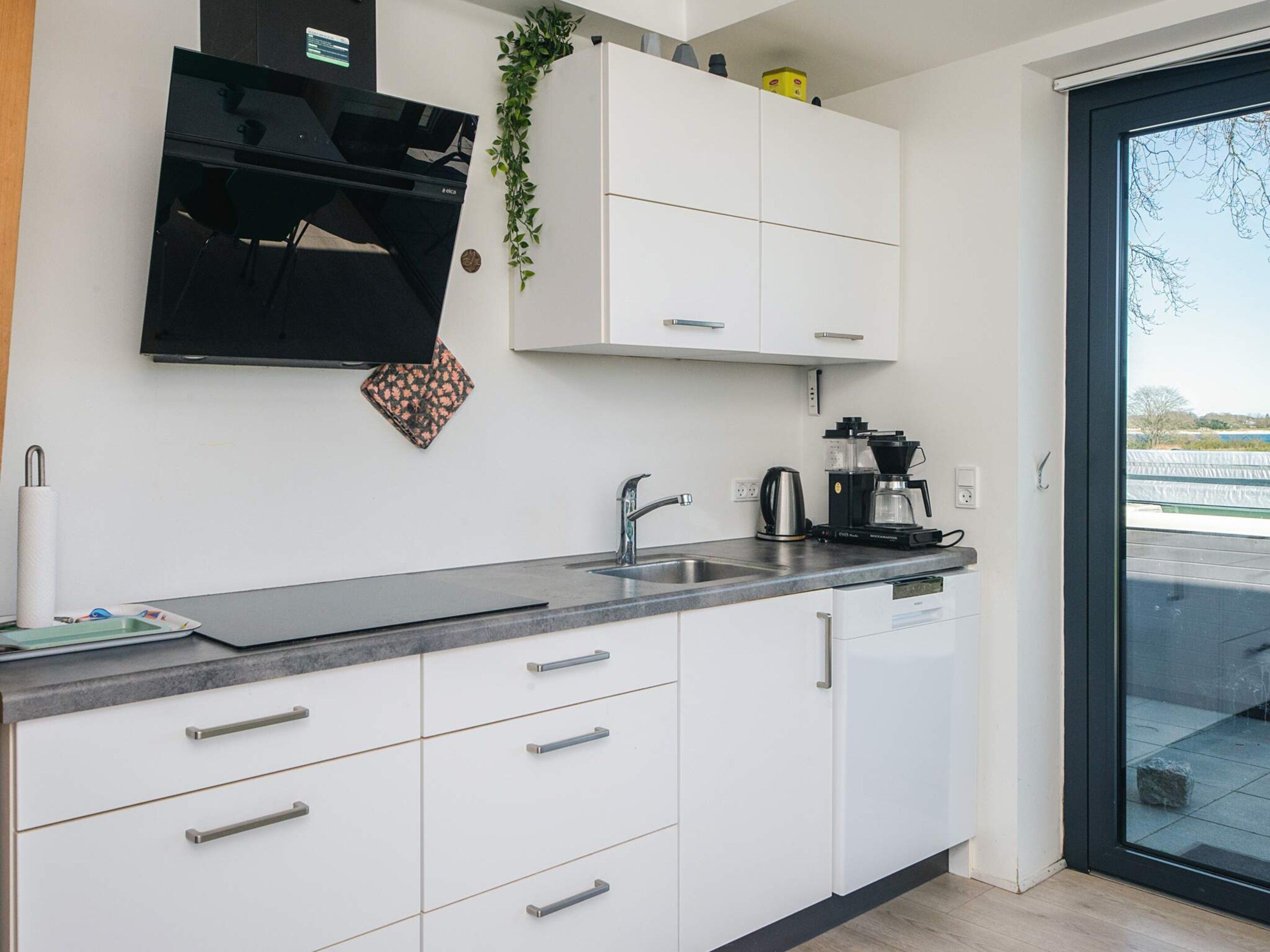 A modern kitchen with white cabinets, a black range hood, a coffee maker, and a view through a glass door.