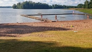 A lakeside beach with a wooden pier extending into the water, a grassy area, and a forested background. - Holiday Home in Gedser (Gamleby)