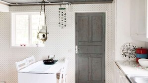 A small kitchen with a white table, chairs, and a wooden floor. There is a window with a hanging light fixture and a door with a calendar on the wall. - Holiday Home in Gedser (Gamleby)