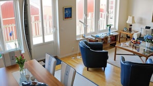 A spacious living room with a wooden dining table, blue armchairs, and a glass coffee table. There are large windows providing natural light and a view of the outside. - Holiday Home in Skärhamn (Skaerhamn)