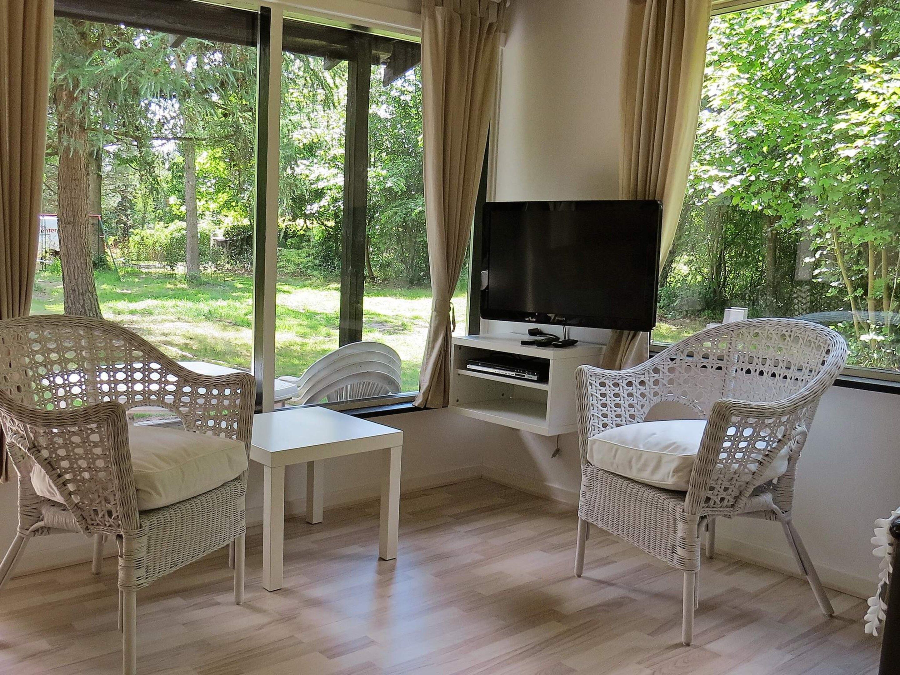 Una habitación con mobiliario de mimbre, un televisor sobre un mueble bajo y vistas a un paisaje verde a través de amplios ventanales.