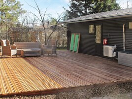 Una terraza de madera con mobiliario exterior, un edificio con puerta verde y un extintor.