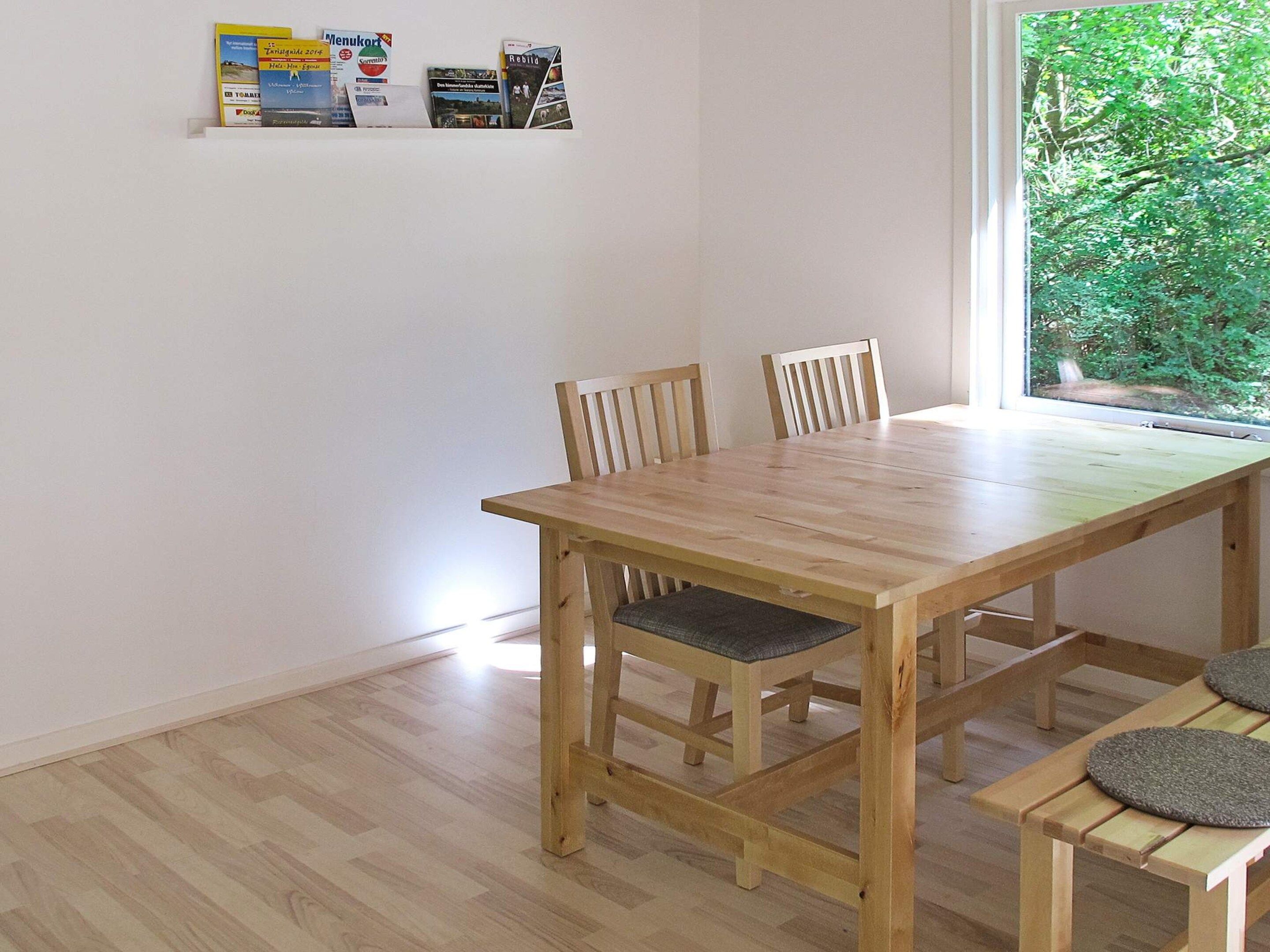 Una mesa de madera para comer con cuatro sillas, un ventanal con vistas a follaje y un estante con varios objetos.